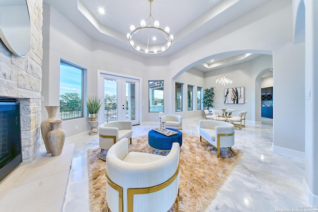 living room with a raised ceiling, french doors, a chandelier, and a stone fireplace