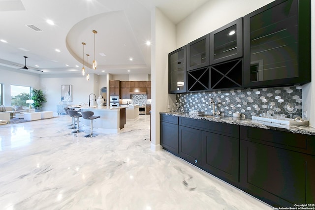 kitchen featuring light stone countertops, a tray ceiling, sink, decorative light fixtures, and tasteful backsplash