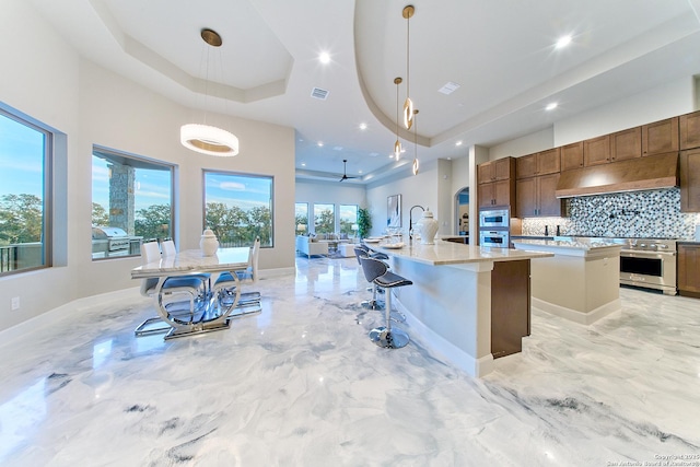 kitchen featuring custom exhaust hood, pendant lighting, a raised ceiling, a kitchen island with sink, and a kitchen breakfast bar