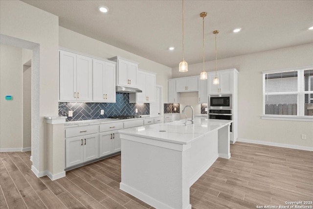 kitchen featuring stainless steel oven, pendant lighting, a center island with sink, built in microwave, and white cabinetry