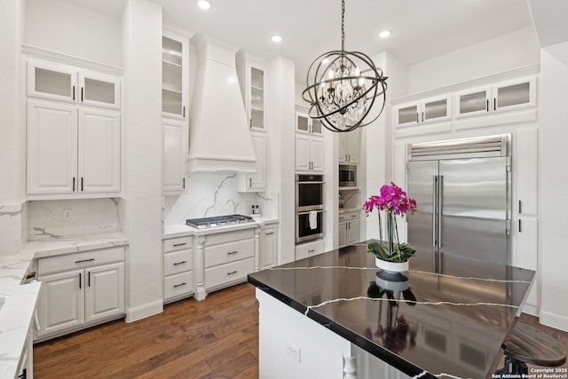 kitchen featuring hanging light fixtures, built in appliances, custom range hood, decorative backsplash, and white cabinetry