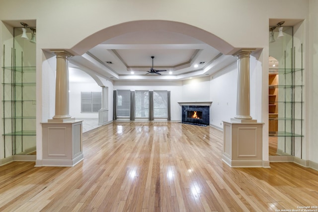 unfurnished living room with ornate columns, light wood-type flooring, ceiling fan, a fireplace, and a tray ceiling