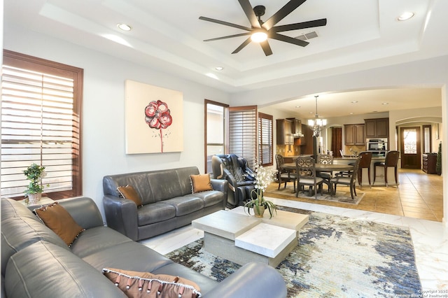 tiled living room featuring a tray ceiling and ceiling fan with notable chandelier