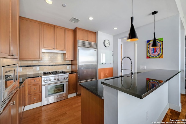 kitchen with pendant lighting, dark stone counters, built in appliances, sink, and light wood-type flooring