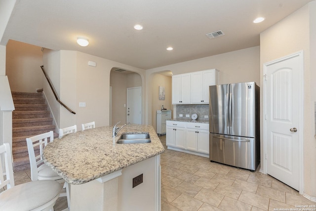 kitchen with light stone countertops, white cabinets, an island with sink, sink, and stainless steel fridge