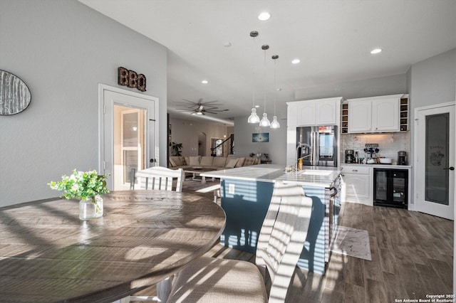 kitchen featuring white cabinetry, stainless steel fridge, an island with sink, decorative light fixtures, and beverage cooler