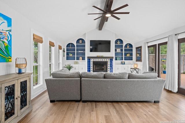 living room with ceiling fan, a tile fireplace, vaulted ceiling with beams, light hardwood / wood-style flooring, and built in shelves