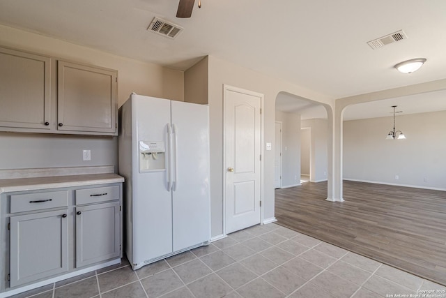 kitchen featuring light tile patterned floors, gray cabinets, hanging light fixtures, white refrigerator with ice dispenser, and ceiling fan with notable chandelier