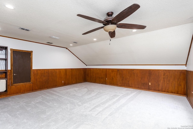 additional living space with a textured ceiling, light carpet, wooden walls, vaulted ceiling, and ceiling fan