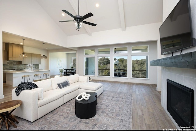 living room featuring light hardwood / wood-style floors, ceiling fan, high vaulted ceiling, beam ceiling, and a tile fireplace