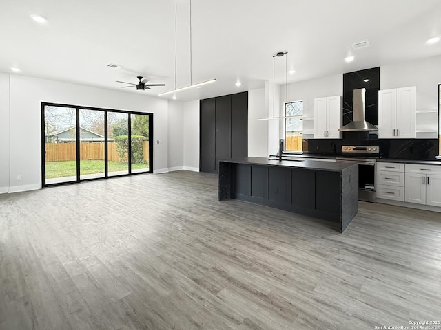 kitchen with stainless steel electric stove, white cabinets, wall chimney exhaust hood, a kitchen island with sink, and ceiling fan