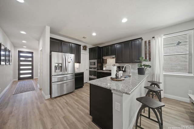 kitchen featuring a breakfast bar area, stainless steel appliances, decorative backsplash, light wood-type flooring, and light stone countertops