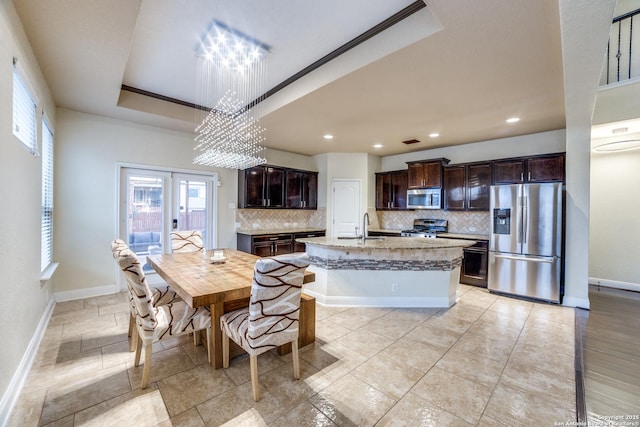 kitchen with appliances with stainless steel finishes, a tray ceiling, a kitchen island with sink, dark brown cabinets, and a chandelier