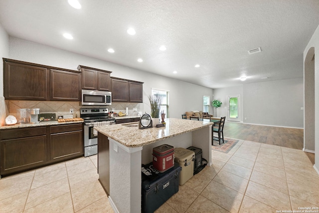 kitchen with appliances with stainless steel finishes, tasteful backsplash, a kitchen island with sink, light tile patterned flooring, and light stone countertops