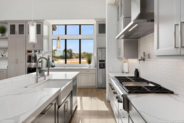 kitchen with light stone countertops, appliances with stainless steel finishes, decorative light fixtures, wall chimney range hood, and light hardwood / wood-style floors