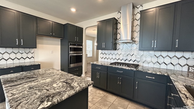 kitchen featuring backsplash, light tile patterned flooring, appliances with stainless steel finishes, wall chimney exhaust hood, and dark stone counters