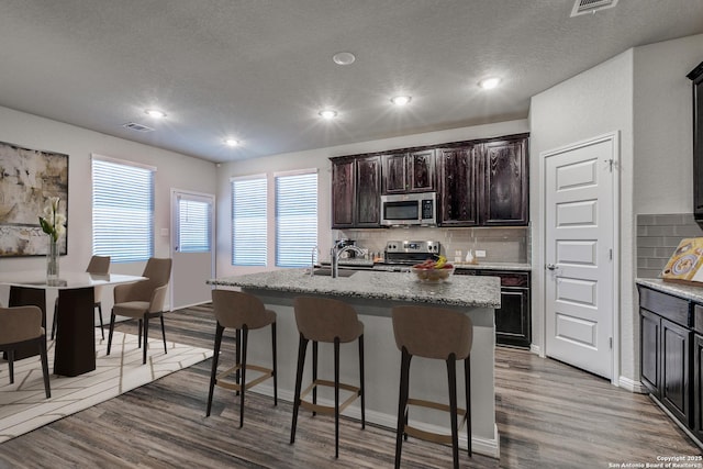 kitchen featuring stainless steel appliances, decorative backsplash, wood-type flooring, and an island with sink