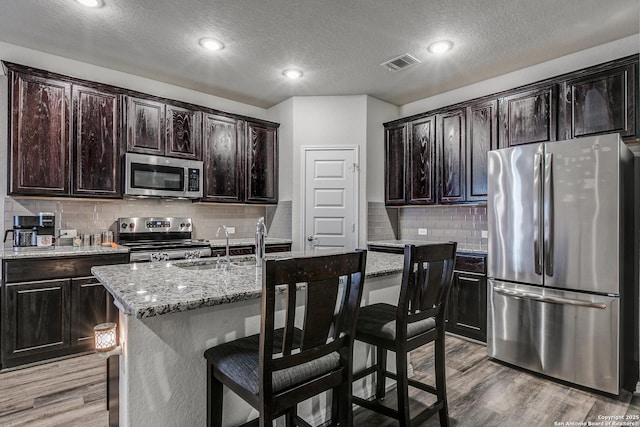 kitchen with stainless steel appliances, a kitchen island with sink, light stone counters, dark brown cabinets, and a breakfast bar area