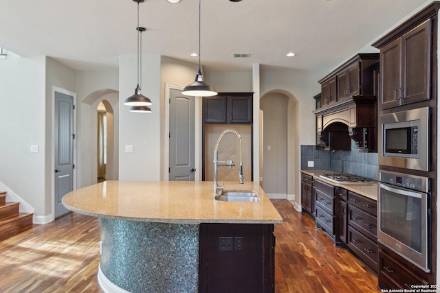 kitchen with decorative light fixtures, backsplash, light stone countertops, appliances with stainless steel finishes, and an island with sink