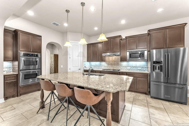 kitchen with sink, hanging light fixtures, a kitchen island with sink, appliances with stainless steel finishes, and light stone counters