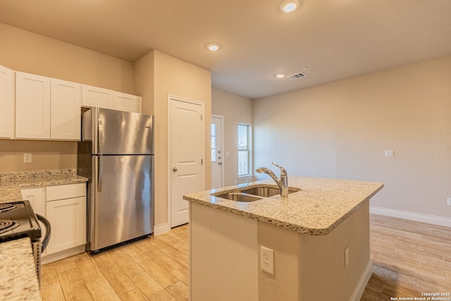 kitchen with light stone countertops, white cabinets, an island with sink, sink, and stainless steel refrigerator