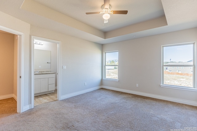 unfurnished bedroom with ceiling fan, ensuite bath, light carpet, and a tray ceiling