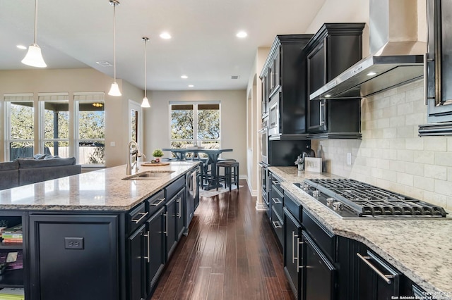 kitchen with wall chimney exhaust hood, decorative light fixtures, sink, stainless steel gas stovetop, and a center island with sink