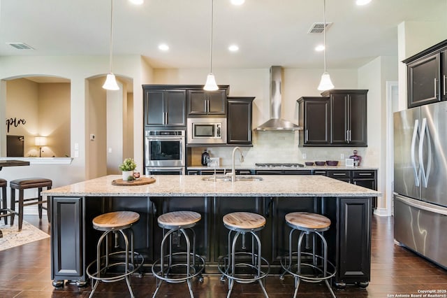 kitchen with pendant lighting, appliances with stainless steel finishes, a center island with sink, and wall chimney exhaust hood