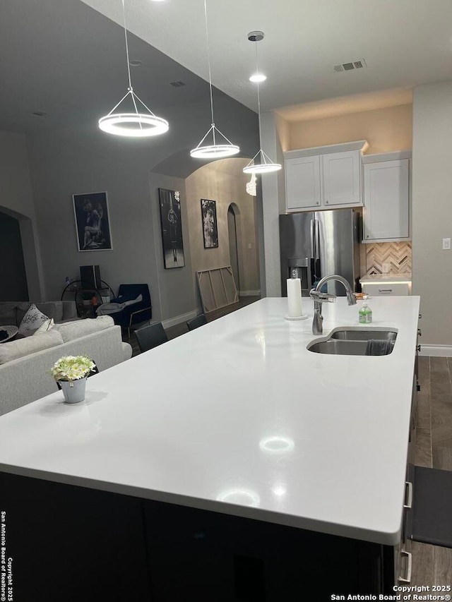 kitchen with sink, stainless steel fridge, white cabinetry, hanging light fixtures, and a center island with sink