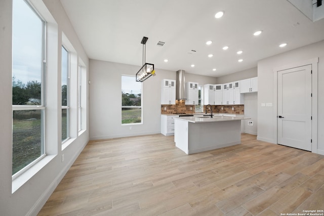 kitchen with decorative light fixtures, wall chimney range hood, a center island with sink, sink, and white cabinets