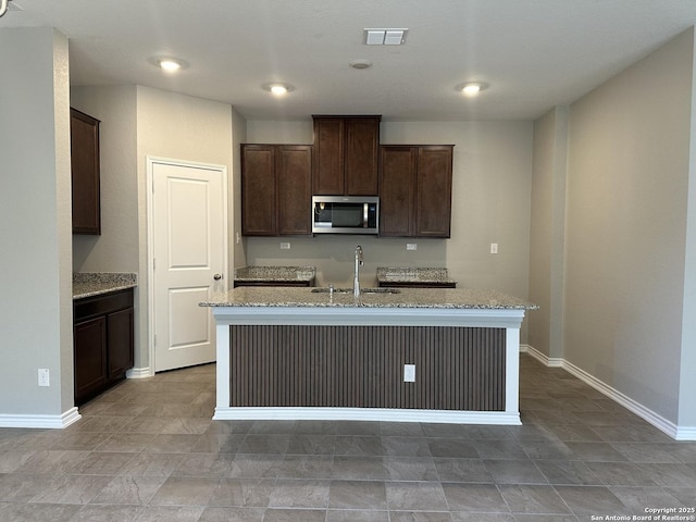 kitchen with light stone countertops, a center island with sink, dark brown cabinetry, and sink