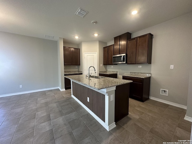 kitchen with dark brown cabinets, a center island with sink, light stone counters, and sink