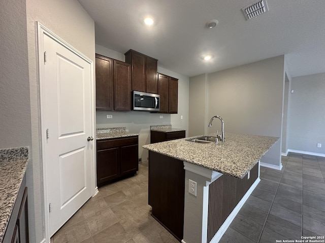 kitchen featuring an island with sink, tile patterned flooring, dark brown cabinets, light stone countertops, and sink