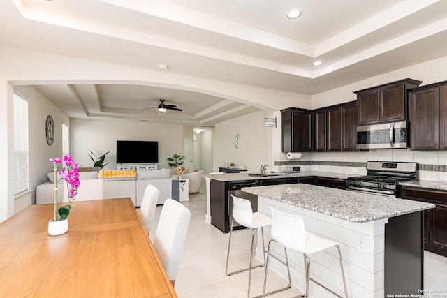 kitchen with ceiling fan, appliances with stainless steel finishes, a tray ceiling, light stone counters, and sink