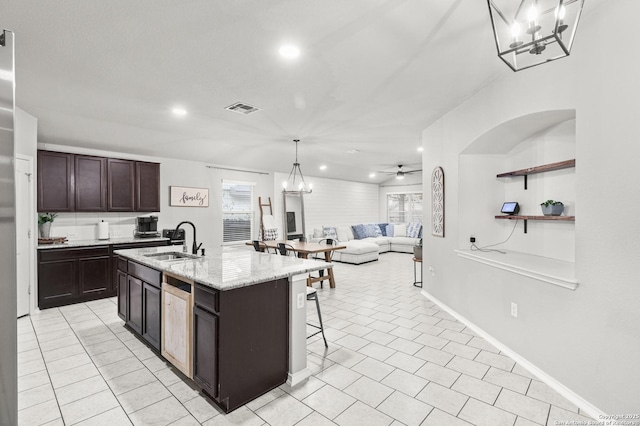 kitchen with light tile patterned floors, a kitchen island with sink, ceiling fan with notable chandelier, dark brown cabinetry, and sink