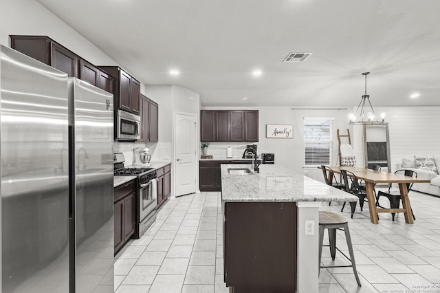 kitchen with an island with sink, stainless steel appliances, decorative backsplash, decorative light fixtures, and light stone countertops