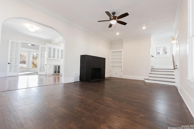 unfurnished living room featuring ceiling fan, a fireplace, dark wood-type flooring, ornamental molding, and built in shelves