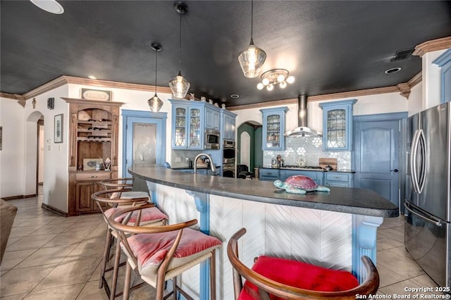 kitchen with light tile patterned floors, arched walkways, dark countertops, wall chimney exhaust hood, and stainless steel appliances