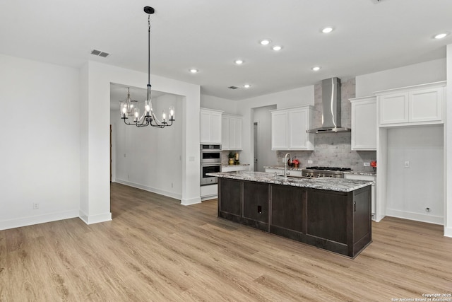 kitchen featuring white cabinetry, a center island with sink, light stone countertops, light hardwood / wood-style flooring, and wall chimney range hood