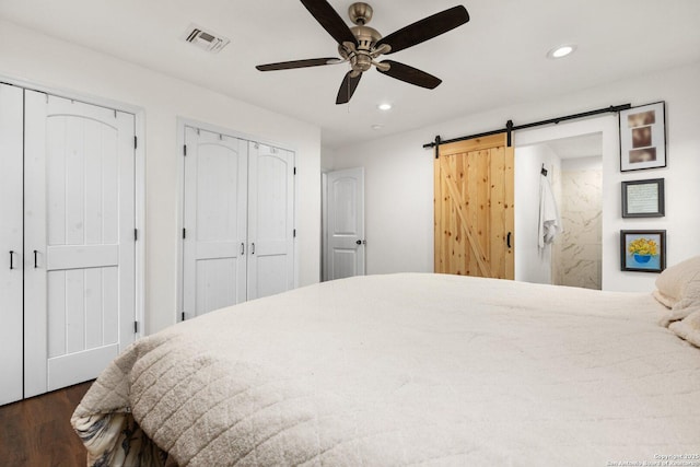 bedroom featuring ceiling fan, a barn door, dark hardwood / wood-style flooring, and two closets