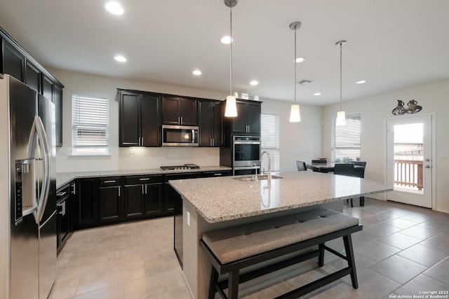 kitchen featuring a breakfast bar area, stainless steel appliances, decorative light fixtures, a kitchen island with sink, and light stone countertops