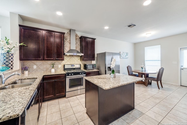 kitchen with light stone countertops, appliances with stainless steel finishes, a kitchen island, wall chimney range hood, and sink