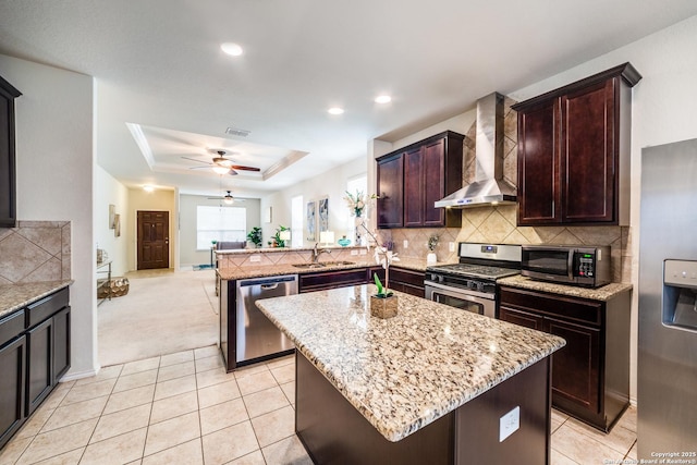 kitchen with appliances with stainless steel finishes, a kitchen island, a raised ceiling, kitchen peninsula, and wall chimney exhaust hood