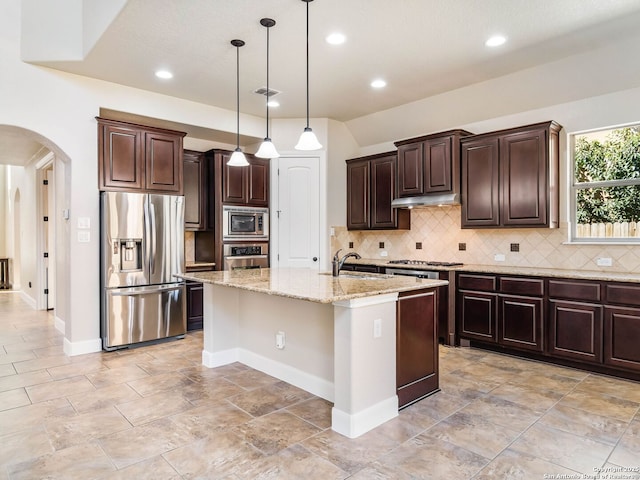 kitchen featuring dark brown cabinetry, hanging light fixtures, a center island with sink, stainless steel appliances, and light stone countertops