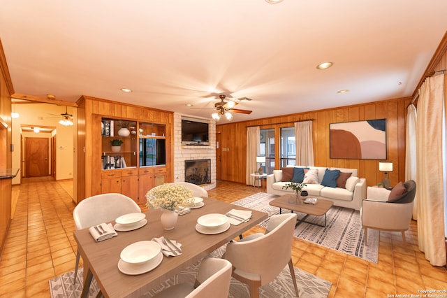 dining space featuring a fireplace, light tile patterned floors, built in shelves, ceiling fan, and wood walls