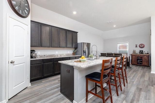 kitchen featuring a kitchen bar, an island with sink, stainless steel fridge with ice dispenser, light wood-type flooring, and light stone counters