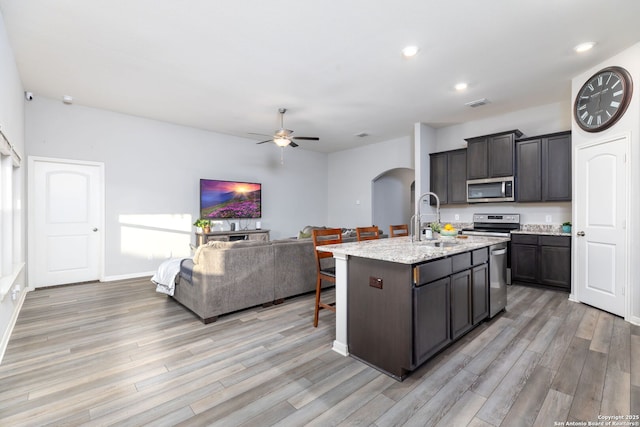 kitchen with light stone countertops, appliances with stainless steel finishes, a kitchen island with sink, sink, and ceiling fan