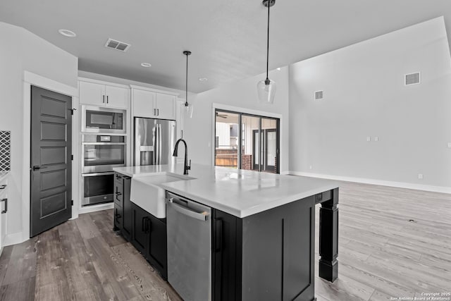 kitchen featuring hanging light fixtures, appliances with stainless steel finishes, white cabinets, light wood-type flooring, and an island with sink