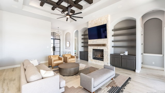 living room with a stone fireplace, built in shelves, ceiling fan with notable chandelier, light hardwood / wood-style floors, and beamed ceiling