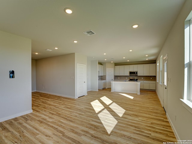 kitchen with an island with sink, light hardwood / wood-style floors, white cabinets, backsplash, and stainless steel appliances